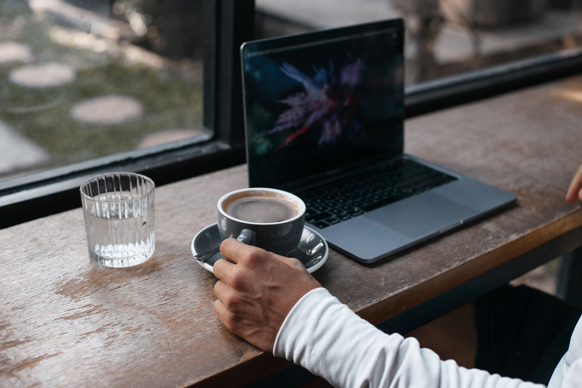 Une main tenant une tasse de café à côté d'un ordinateur portable posé sur une table en bois, près d'une fenêtre.