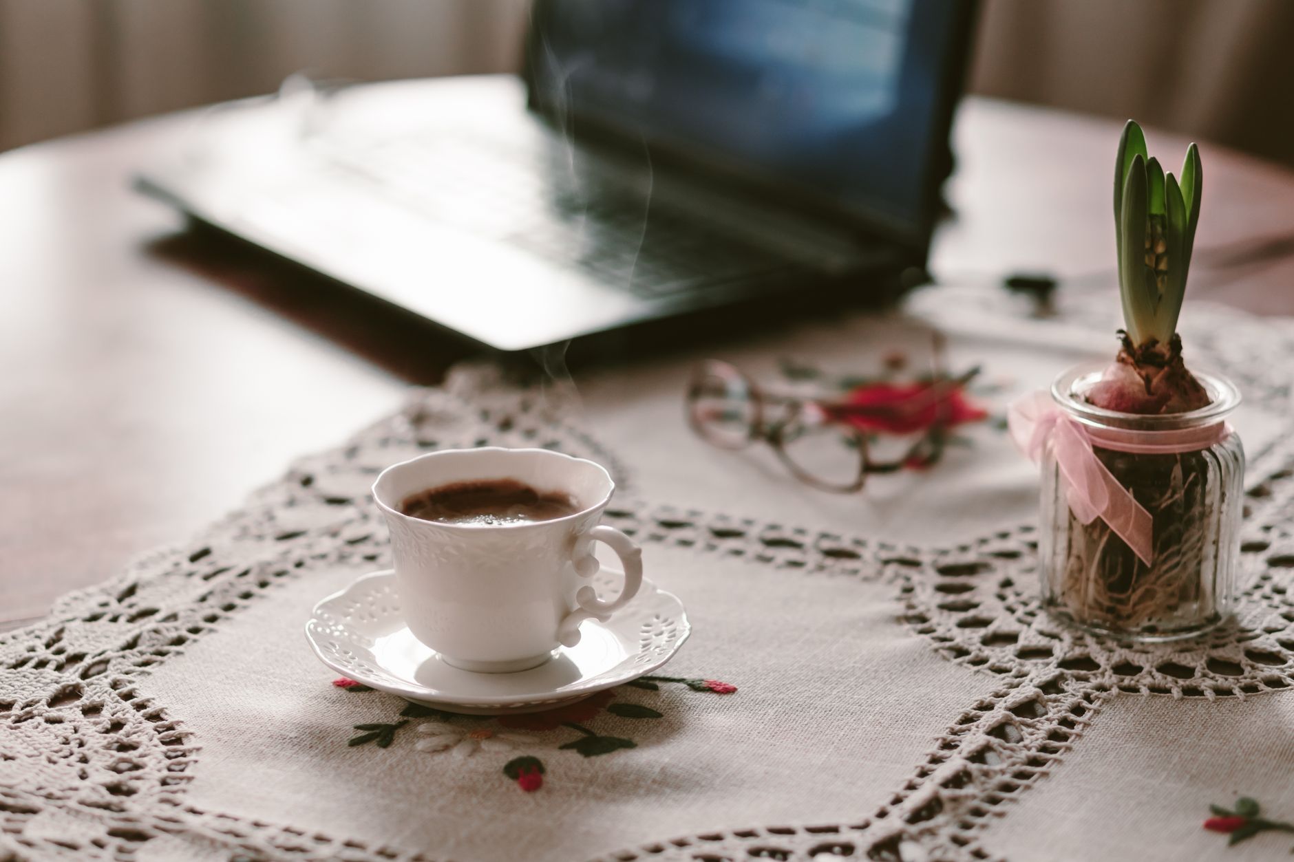 Un espace de travail chaleureux avec une tasse de café fumante sur une nappe en dentelle, à côté d'un ordinateur portable.