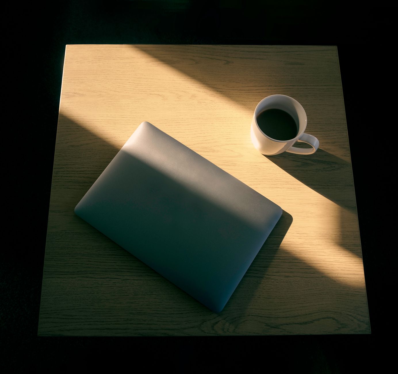 Une installation minimaliste avec un ordinateur portable et une tasse de café sur une table en bois, jeu d'ombre et de lumière.